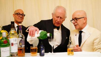 King wearing black tuxedo pours alcohol from bottle into glass as two men in suits watch on