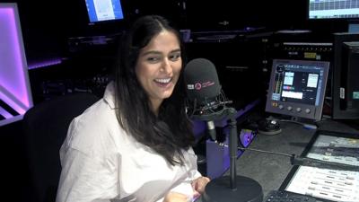 Gura smiles in front of an Asian Network mic in the studio, wearing a white shirt.