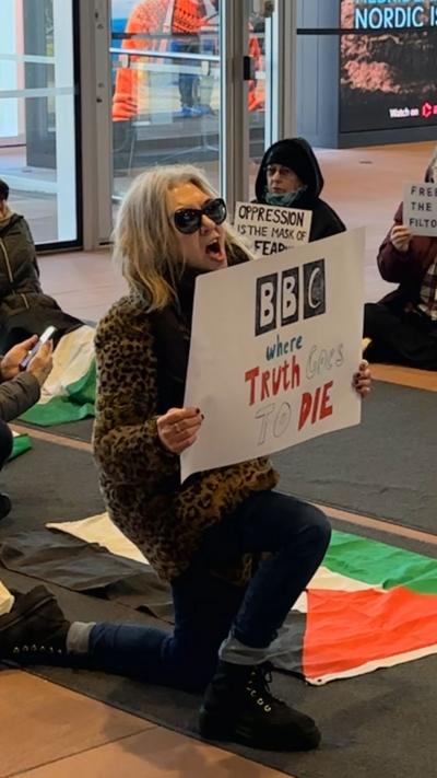 Protesters insid BBC Scotland HQ reception. There is a woman at the front kneeling down holding a protest sign with a Palestinian flag lying on the floor next to her. There are five other people sitting on the ground around her also holding protest signs.