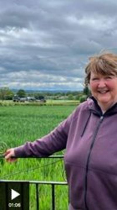 A woman stands smiling at the camera. She has short light brown hair and is wearing a purple zip up fleece. Behind her is a cloudy sky, green field, and a gate which she is leaning on.