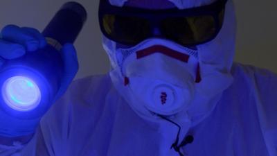 Jim Gildea is shown in full PPE, holding a torch emitting a blue light. He wears tinted lenses over his protective glasses as he looks for evidence of any remaining biological matter.
