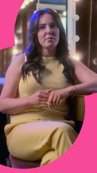 A woman with long brown hair sits in a chair. She wears a yellow sleeveless top and yellow trousers 