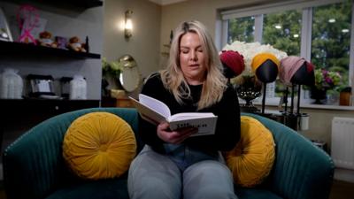 Ashleigh Palmer reading a book. She is sitting on a dark green couch which has two round, yellow cushions on it. She has long blonde hair and wears a black round neck top and light blue jeans. Behind her are shelves containing items including teddy bears. There is a wooden desk with an oval mirror on it, a table with large white flowers and a white uPVC window made up of three sections and topped with a white roller blind that is fully open. Large, lush green trees can be seen outside.