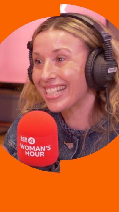 Kate Weir sits in-front of a red BBC Radio 4 Woman's Hour mic and smiles. She is wearing black headphones. 