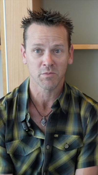 Man seated looking into the camera wearing a green plaid button up shirt. 