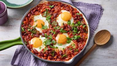 A pan with a green handle containing beans and four eggs. It sits on a purple cloth with a wooden spoon by the side