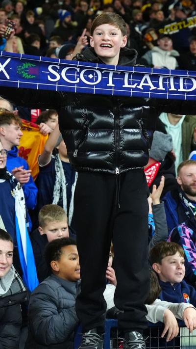 Young boy standing among a crowd of football fans, holding a scarf that reads "Scotland"