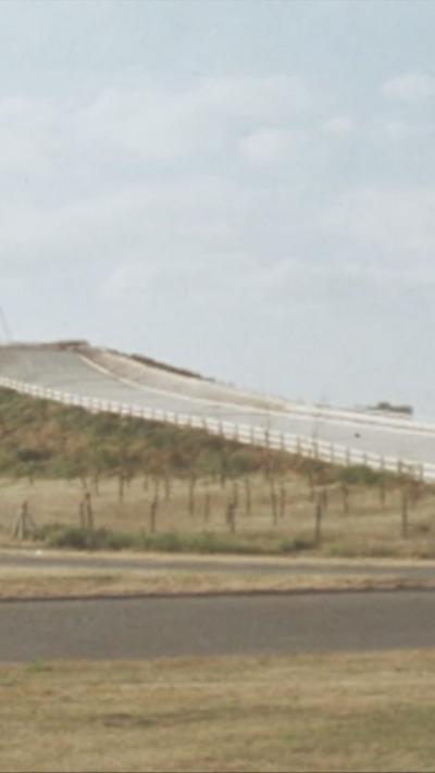 A large, grey ski slope located right on top of a hill and by the side of the motorway