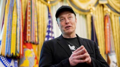Elon Musk, wearing black, with hands together, fingertips touching, and a baseball cap.