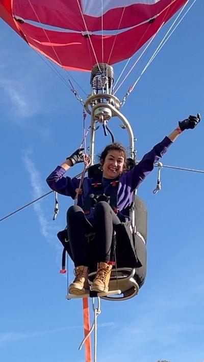 Woman seated below a hot air balloon, celebrating with a hand in the air.
