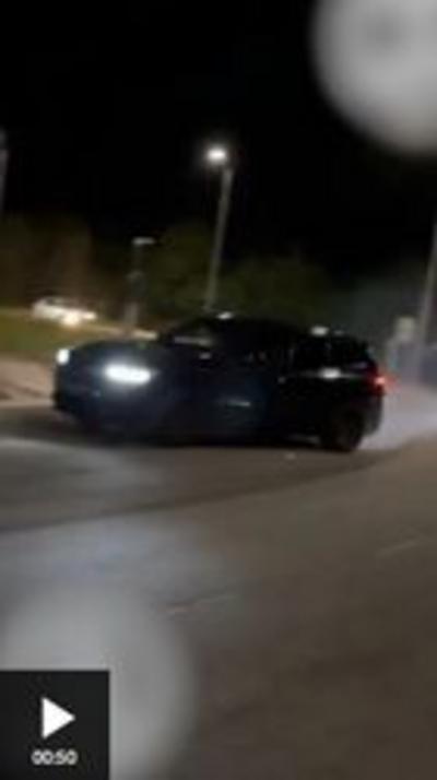 A black van drifting around a roundabout at night. The road ir blurred, there is a streetlamp in the backgroud, and smoke can be seen coming from the van's rear tyres.