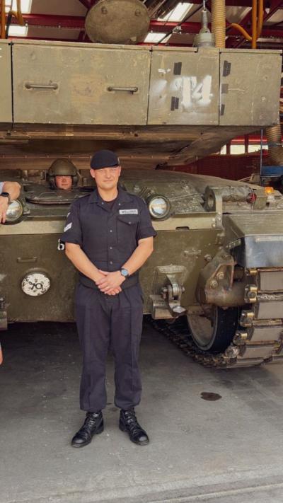 A man wearing a black military uniform, standing in front of a large military tank.