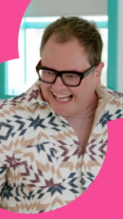 Alan Carr smiles with his hands outstretched. He wears black rimmed glasses and a patterned shirt