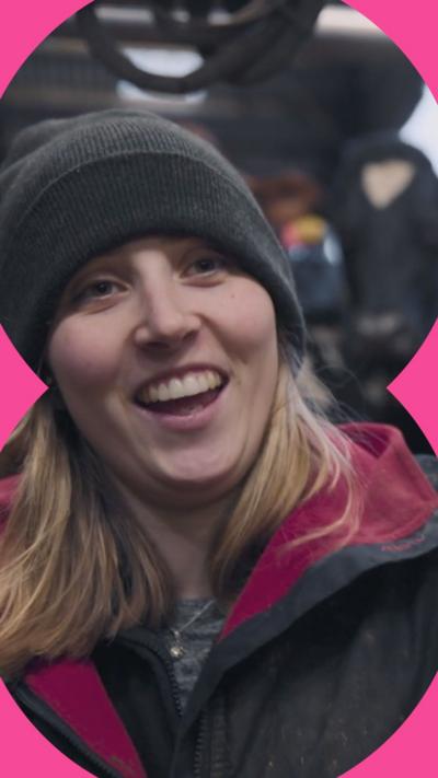 A female farmer in a shed full of cows smiles happily at the camera