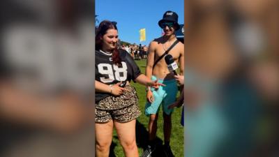 A woman and man smiling at TRNSMT festival - the woman is posing with index fingers pointing left, the man has his top off.