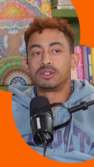 Jordan Stephens is wearing a light blue hoodie and sitting behind a microphone. He has short, curly brown hair.