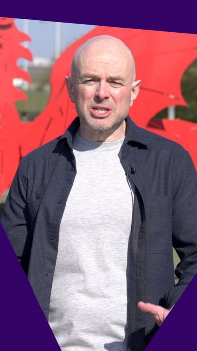 Gareth Lewis speaks to the camera on a sunny day, in the background there is a sculpture of a red dragon.
