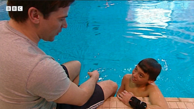 Gethin Jones on the left hand side, sitting on the edge of the side of a swimming pool.  Tom Daley is in the pool on the right hand side, leaning on the edge looking up at Gethin.