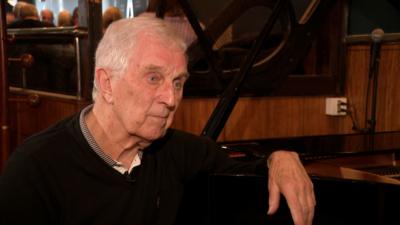 Bill Finlayson sits and rests his arm on a baby grand piano in a bar