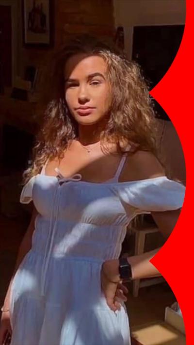 Brooke George is looking at the camera wearing a cotton off-the-shoulder dress. She has long wavy blonde hair and is posing for the camera with her hand on her hip
