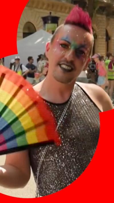 A man with pink hair and face paint waving a rainbow fan and smiling