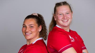 Standing back-to-back with their arms crossed, sisters Gwenllian Prys and Alaw Pyrs, who is slightly taller, both in red Wales rugby kits, smile into the camera against a plain white background
