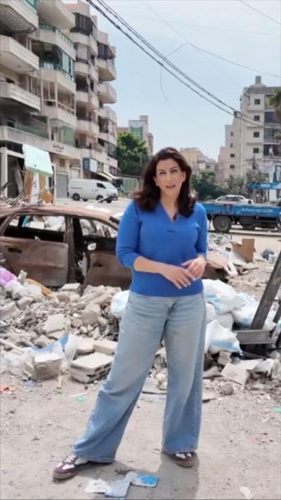 Carine Torbey in a blue top and jeans, stood next to rubble and a destroyed car