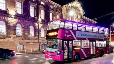 A purple double-decker bus drives past a large cream building at night. 