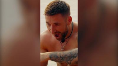 A topless man looks stunned. He has tattoos and a necklace.