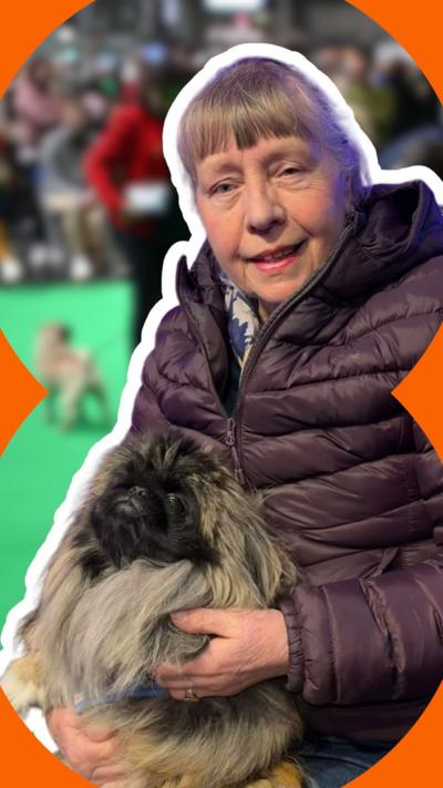 Eileen sits with a small brindle-coloured dog on her knee. She has grey hair pulled back from her face with a full fringe. She wears a brown puffer jacket.