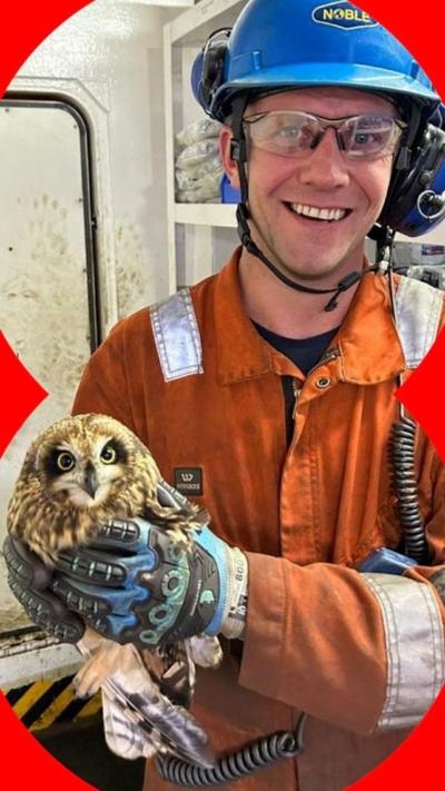 A man wearing an orange oil rig uniform and blue hard hat, smiles to camera, holding an owl with both hands.