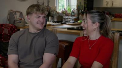 Race Across the World contestants Fin and Sioned smile at eachother inside a kitchen. 