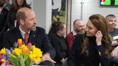 Prince William and Princess Catherine engaged in conversation at a table. He has his hand out and she is holding her left ear. A vase of flowers is visible on the table in front of them and people are talking and smiling in the background