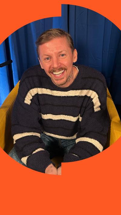 Professor Green smiling to camera. He wearing a navy and white stripy jumper.