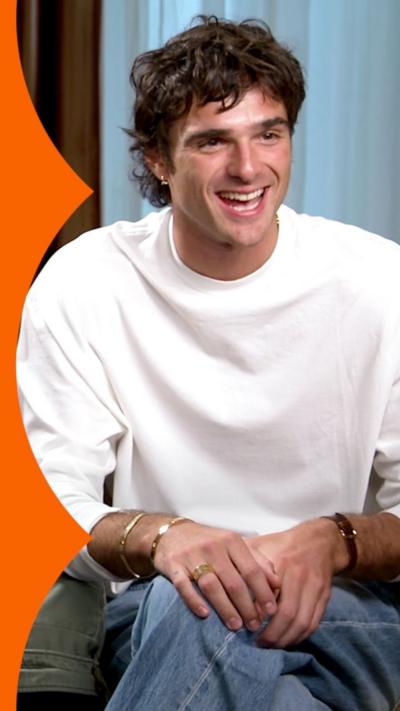 A man, Jacob Elordi, has dark brown hair and wears a white top. He is smiling.