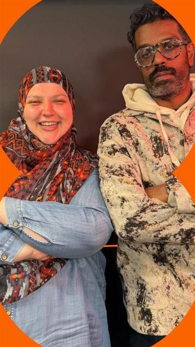 A woman wearing an orange, patterned headscarf smiles at the camera. She stands next to a man with short, curly hair wearing glasses. He looks at the camera. They both have their arms folded.