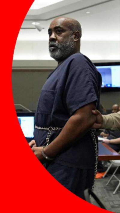 A man with a grey beard and is wearing a blue prison jumpsuit and handcuffs.
