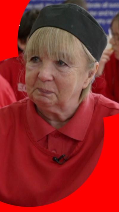 A dinner lady with blonde hair smiles at someone off camera. She is wear a black uniform cap, a red T-shirt with a button-up collar, and red overalls.