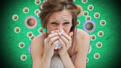 A woman blows her nose in front of a diagram featuring diagrams of viruses