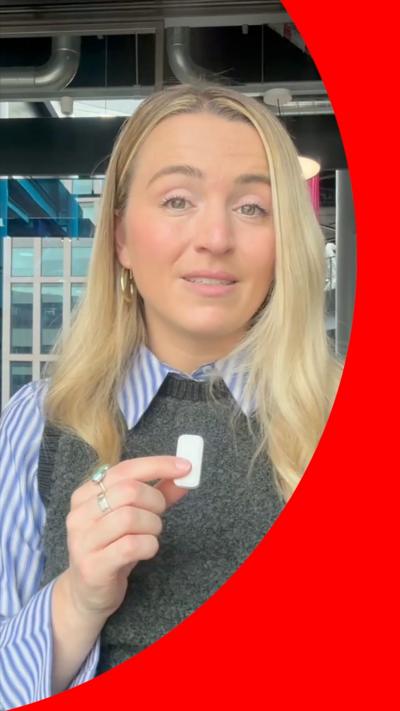 BBC Journalist Lucy Woodham wears a blue striped shirt with a grey pullover and looks at the camera. She holds a small white microphone with her right hand.