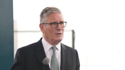 Prime Minister Sir Keir Starmer, he had short, grey hair and is wearing glasses, a dark suit and green tie.