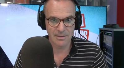 Martin Lewis wears black shirt with white stripes and glasses. He sits behind a microphone