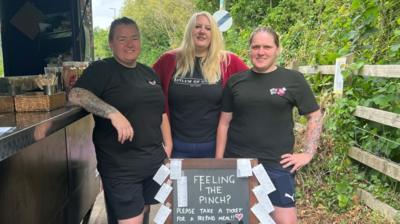 Three women stand in front of an advertising board with feeling the pinch written on it