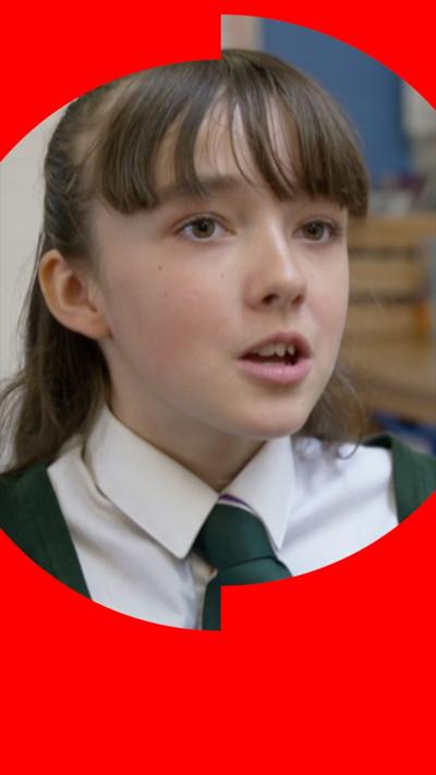 A girl with brown hair and a fringe is looking off-camera. She is wearing a white shirt, and green school tie and blazer.