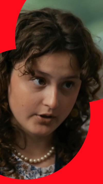Close-up of a young person with curly hair, wearing a pearl necklace and patterned top.
