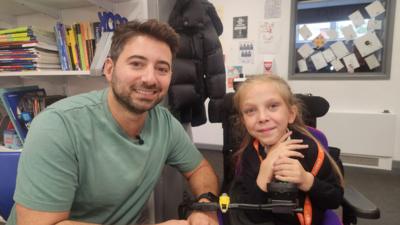 Ricky sitting next to Isla, who is in a wheelchair next to him. Both are smiling.