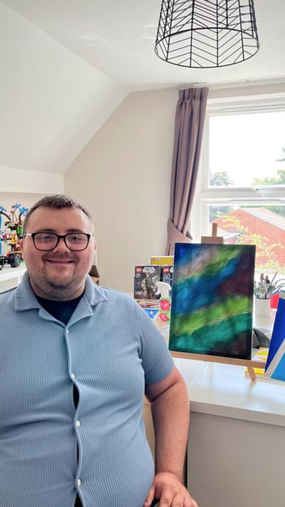 A man with glasses and beard wearing a blue shirt sat in an art studio