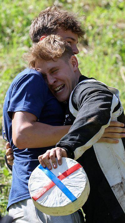 Two men hugging as one of them holds the winning cheese wheel.