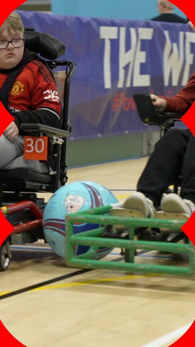 A blue football is wedged between two powerchairs being driven in a sports hall. A young boy is seen on the left wearing a Manchester United FC jersey.