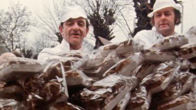 Two men in baker's uniforms stood behind a huge pile of hot cross buns on a market stall. 